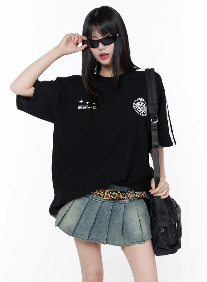 LEWKIN Oversized Athletic Tee CU519 in black (M/L), front three-quarter on model wearing sunglasses, drop-shoulder with white sleeve stripes and eagle crest graphic, styled with pleated denim mini skirt, leopard belt, and black crossbody bag.