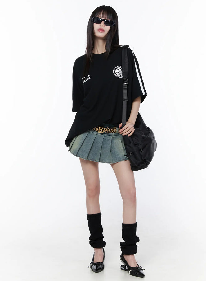 Pleated Denim Mini Skirt CU519 in Blue, full-body front styling shot, worn with oversized black graphic tee, leopard belt, black leg warmers, tote bag and sunglasses.