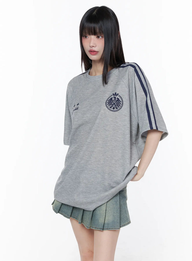 Oversized Athletic Tee CU519 in grey, front view on model, drop-shoulder silhouette with navy sleeve stripes and embroidered eagle crest, paired with pleated denim mini skirt, relaxed athletic fit.